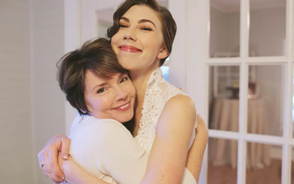 A smiling adult daughter hugs her mother from behind in a bright room with glass doors