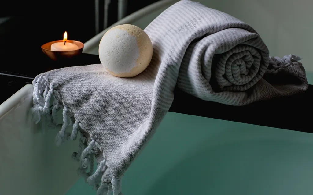 A bath bomb resting on a folded gray towel on the edge of a bathtub next to a lit candle
