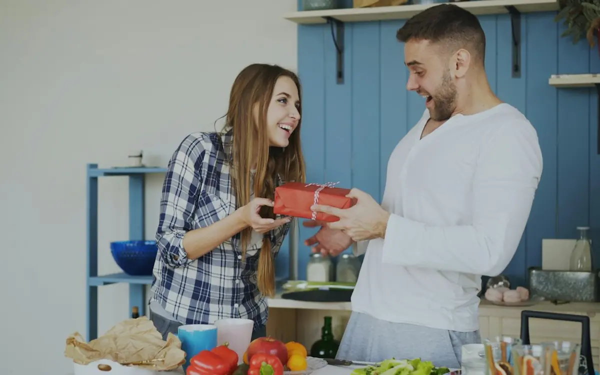 A woman giving her boyfriend a wrapped birthday gift in the kitchen