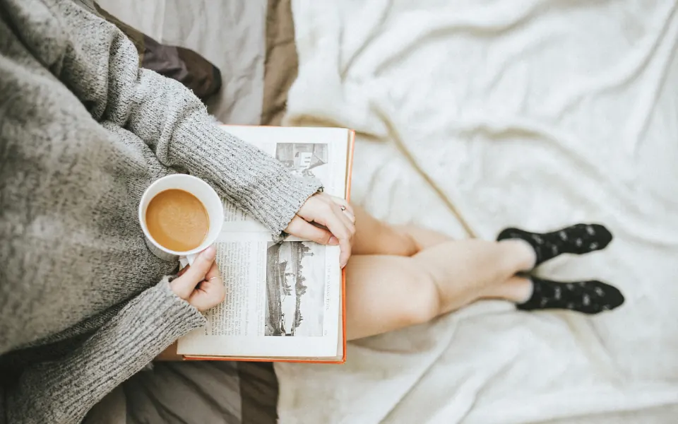 A woman in a gray sweater reading a book in bed with a cup of coffee