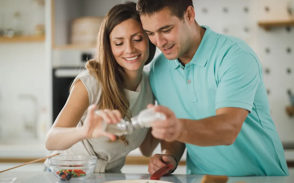 A smiling couple cooking together in a bright kitchen, adding seasoning to a dish