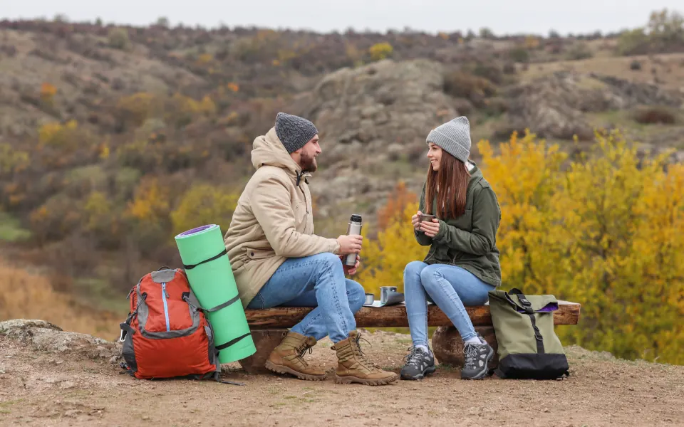 A couple in hats sharing a hot drink from a thermos on a fall hike