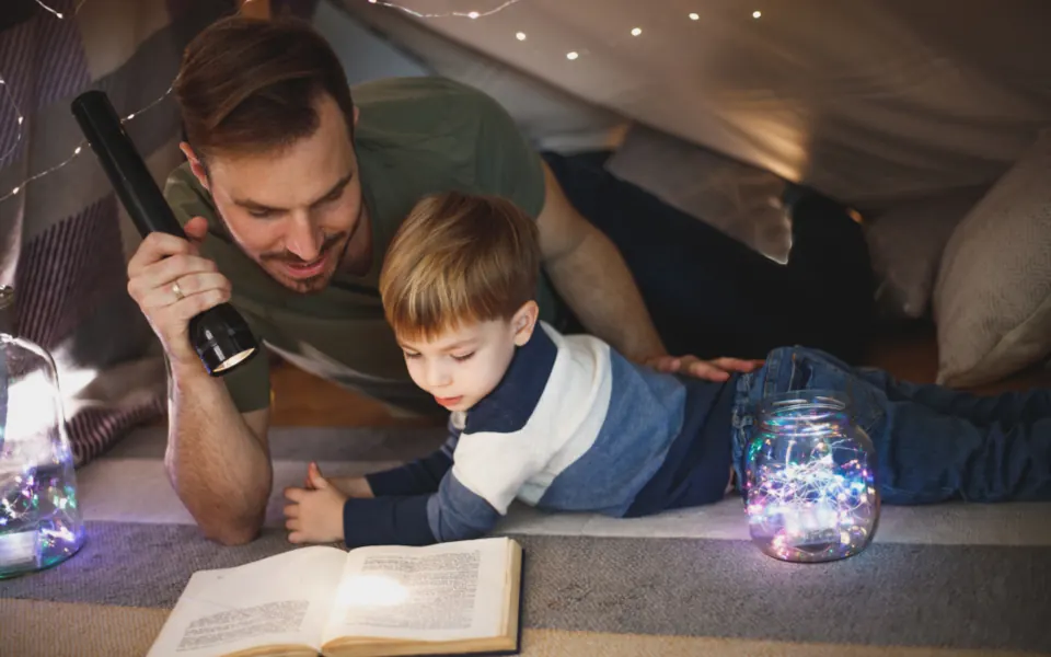 A dad holds a flashlight while reading a book with his young son inside a blanket fort lit with fairy lights