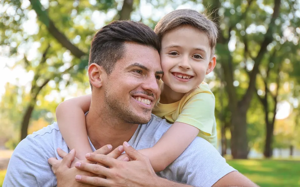 A smiling young boy hugs his dad from behind in a sunny park