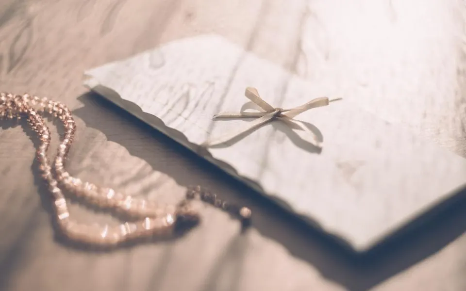 A white envelope tied with a thin ribbon bow lying on a wooden table next to a pearl necklace