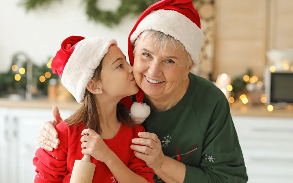 A young girl in a Santa hat kisses her smiling grandmother on the cheek in a kitchen decorated for Christmas