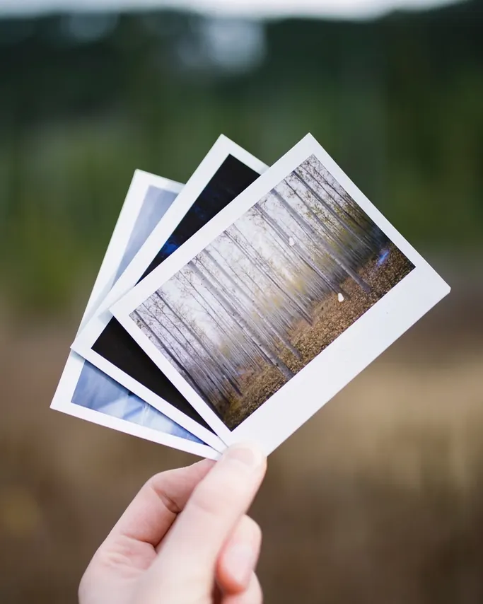 A hand holding up three instant photos outdoors with green trees blurred behind