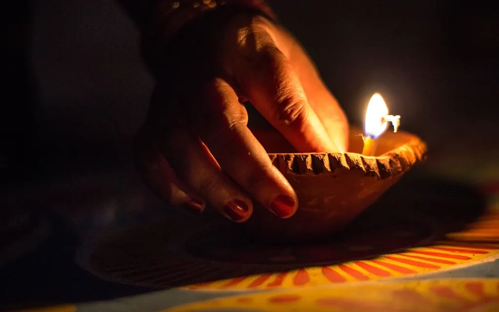 A hand placing a lit clay diya on a colorful rangoli in the dark