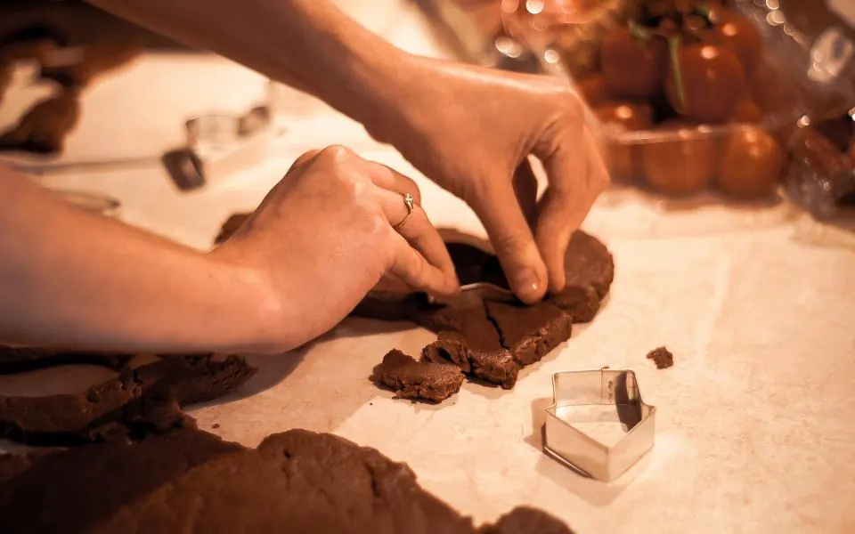 Hands pressing cookie cutters into dark gingerbread dough on a floured counter in warm light
