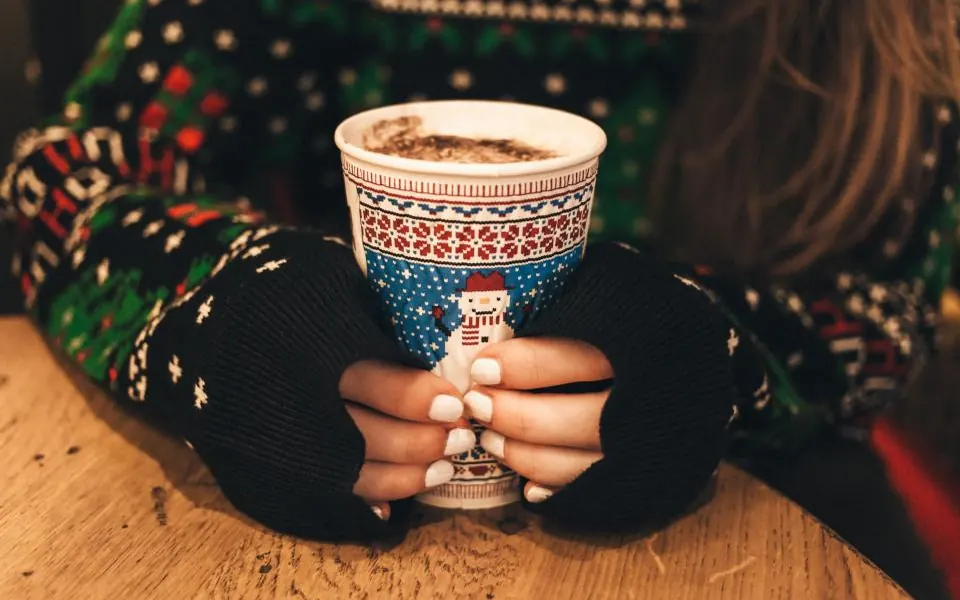 Hands in black fingerless gloves holding a holiday cup with a snowman pattern, with a Christmas sweater behind