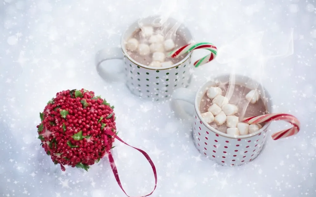 Two mugs of hot cocoa with marshmallows and candy canes next to a red berry ornament in the snow