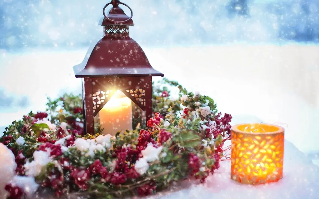 A red metal lantern with a glowing candle inside, sitting in a snowy wreath of red berries next to a small candle holder