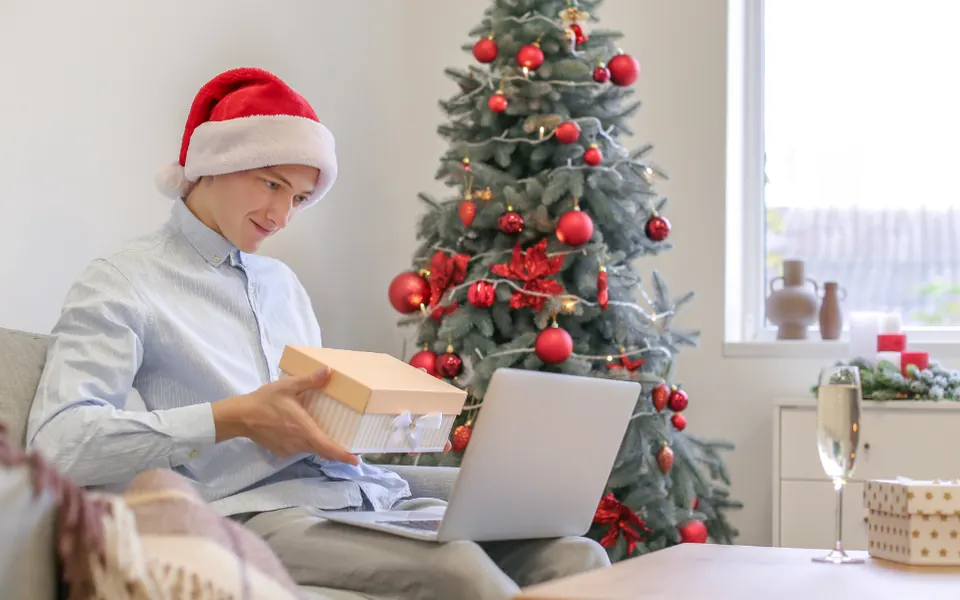 A man in a Santa hat opening a small gift box on the couch next to a decorated Christmas tree