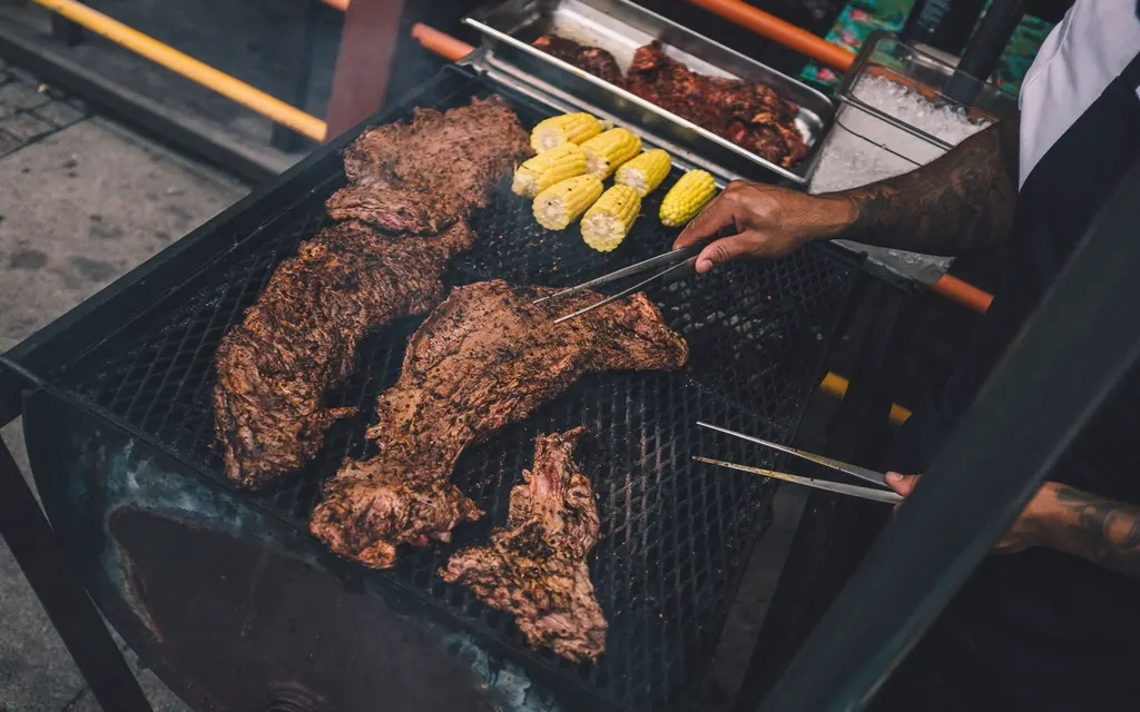 Thick steaks and corn on the cob cooking on a charcoal grill while a hand turns the meat with tongs