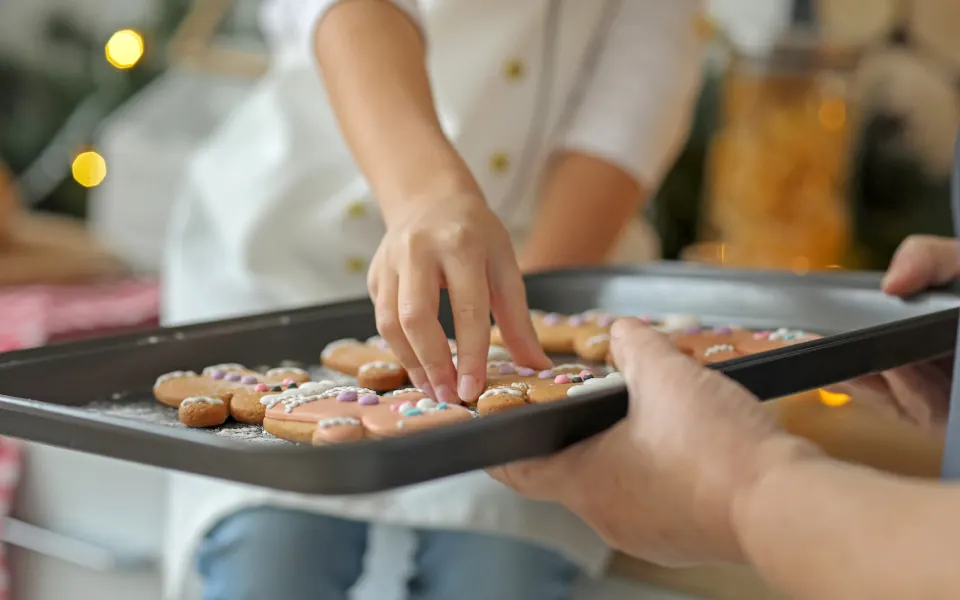 A child reaches for a decorated gingerbread cookie on a baking tray held by an adult, with Christmas lights in the background