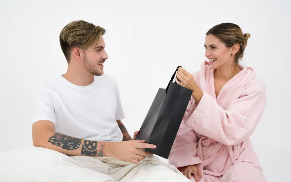A smiling woman in a pink robe handing a black gift bag to her husband in bed
