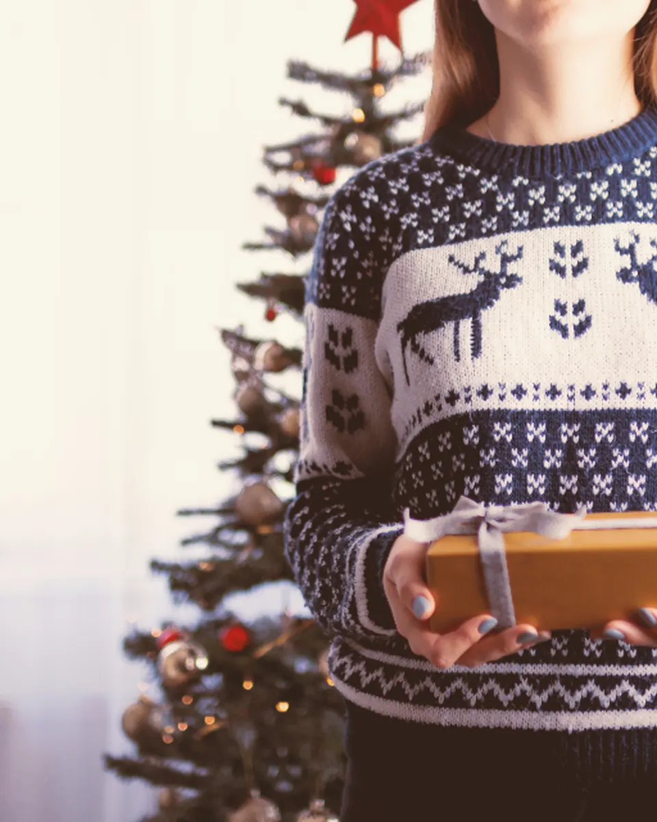 A woman in a navy reindeer sweater holding a small wrapped gift in front of a Christmas tree
