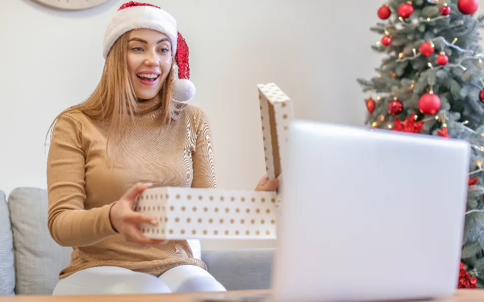 A smiling woman in a Santa hat opens a gift box in front of a laptop, with a decorated Christmas tree behind her