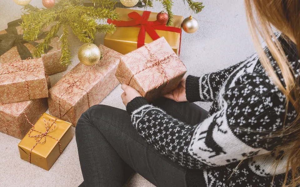 A woman in a Christmas sweater sitting on the floor next to wrapped presents under the tree